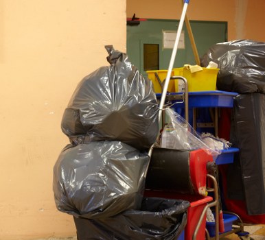 The image shows a collection of black rubbish bags made from durable plastic, stacked on a red trolley with a black cylindrical container placed on top. The rubbish bags appear to be filled with waste and are positioned against a pale yellow wall, with some bags leaning on each other in a seemingly organized manner. To the right, additional black rubbish bags are visible, also placed on or near the trolley. Behind the trolley, there is a small blue shelving unit holding a yellow bin and a white mop with a yellow handle, which is partially visible. The setting appears to be a storage or back area, possibly in a commercial or residential context, within a Greenwich location, consistent with waste collection activities managed by Commercial Waste Removal Greenwich. The environment suggests an indoor or enclosed space designated for waste storage before pickup and removal, with adequate lighting and a utilitarian appearance fitting a typical rubbish holding area.