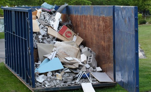 A large industrial skip container painted in faded blue, positioned outdoors on a grassy area near a paved surface, filled with a mix of construction and renovation waste including broken drywall, plasterboard, concrete debris, grey and white insulation boards, and scattered cardboard boxes, some with visible labels and logos. The waste appears to be loosely piled, with materials overlapping and spilling slightly over the top edges of the skip. The interior of the skip reveals a rusted metallic surface, indicating frequent usage over time. Surrounding the container, there are trees and greenery in the background, suggesting the waste is collected in a suburban or urban neighbourhood. The overall scene depicts an active rubbish removal process, possibly carried out by Commercial Waste Removal Greenwich, with a focus on disposing of building refuse, typical of waste management services provided in Greenwich and the surrounding areas of South East London.