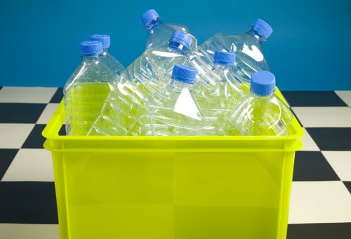 A bright yellow plastic bin filled with several clear plastic bottles with blue caps, some of which are slightly crushed or bent. The bottles are arranged in a haphazard manner, with a few leaning against the edges of the bin. The bin is placed on a black and white checkered surface, likely a floor or tabletop, with a plain blue background behind it. This setup suggests a focus on small plastic waste, commonly handled by rubbish removal services in Greenwich offering waste collection for recyclable materials. The scene conveys a clean, organized approach to waste separation, aligning with professional waste management practices in the local area. The lighting is even, with no shadows obscuring the details of the bottles or the bin, allowing clear visibility of the plastic packaging, textures, and the overall arrangement, making it suitable for a waste removal service website like commercialwasteremovalgreenwich.co.uk specializing in rubbish collection.