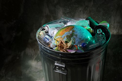 A closed metal rubbish bin filled with various types of discarded waste, including crumpled brown paper, several plastic bottles of different sizes and colors, and a small globe model with visible landmasses and oceans, all surrounding the waste items. The bin is situated outdoors on a paved surface, with a dark, uneven concrete or stone wall in the background. The metallic surface of the bin has a reflective finish, highlighting the surrounding environment under diffuse lighting conditions. This image relates to waste management and rubbish collection services, such as those provided by Commercial Waste Removal Greenwich, and exemplifies typical refuse found during rubbish removal tasks in urban areas near Greenwich or surrounding postcode regions.