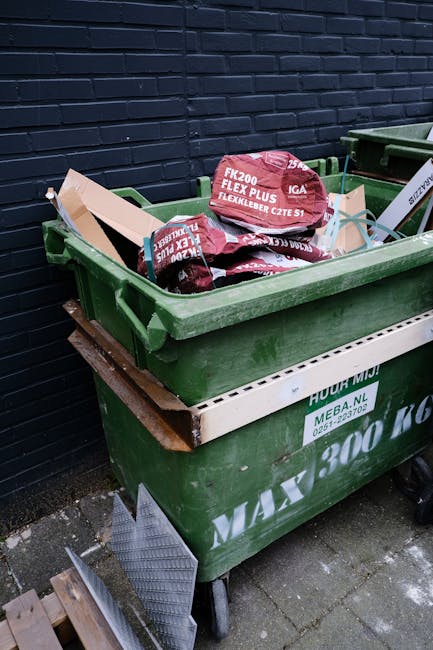 A large green wheeled dumpster filled with various types of waste, including pink and red bags of building materials labeled 'FLEXLEIBER CZTE S1' and other construction debris, positioned outdoors aga