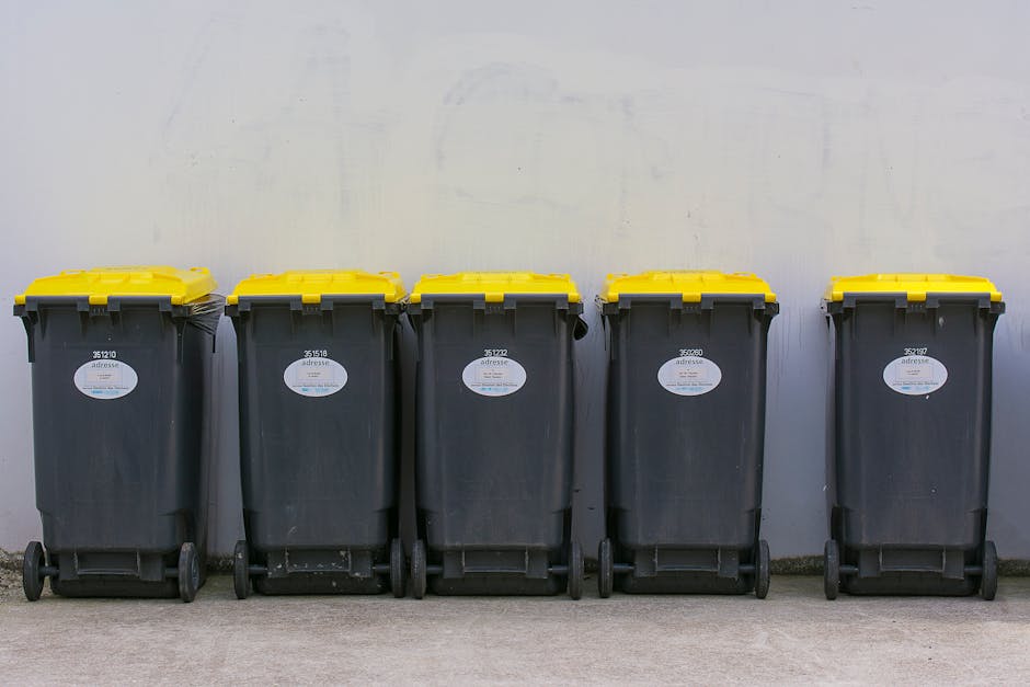 A large green wheeled dumpster filled with various types of waste, including pink and red bags of building materials labeled 'FLEXLEIBER CZTE S1' and other construction debris, positioned outdoors aga