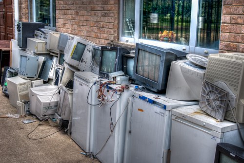 A row of discarded outdated computer monitors and CRT televisions lined up outside against a brick wall of a building, with some showing visible cables and wires hanging loose. The monitors vary in size and color, predominantly in shades of gray, black, and beige, with textured plastic casings. They are positioned on top of several white appliances, possibly old refrigerators or freezers, on a paved surface next to a window reflecting greenery. The arrangement appears to be part of a waste collection effort for electronic waste, possibly as part of rubbish removal services in Greenwich. The setting suggests an exterior location adjacent to a commercial or residential property, and the image highlights the need for professional disposal of bulky electronic waste in accordance with local waste management regulations.